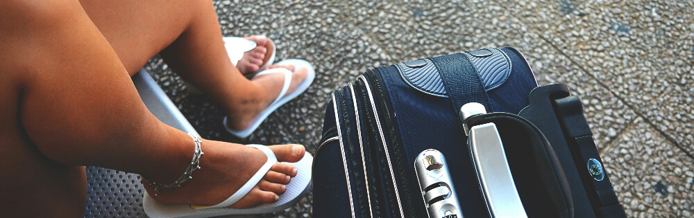 Person sitzt auf einer Bank mit einem schwarzen Reisekoffer daneben und trgt weie Flip-Flops sowie ein Fukettchen.
