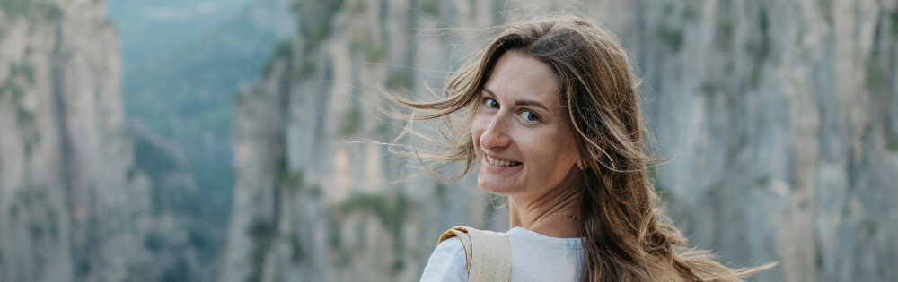 Frau mit langen Haaren, die ins Bild lchelt; im Hintergrund sind steile, felsige Klippen und Natur zu sehen.