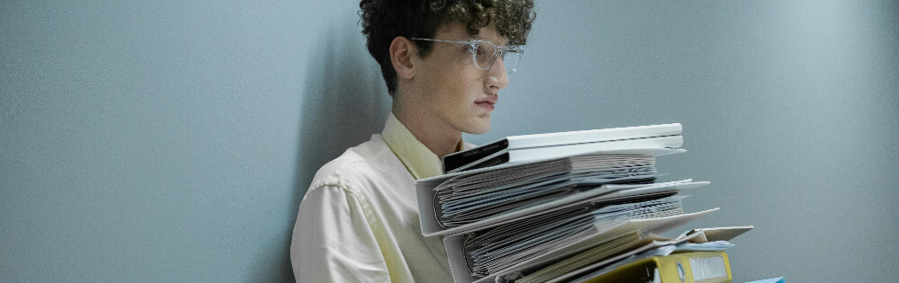 Ein junger Mann mit Locken und Brille steht an einer grauen Wand und hlt einen groen Stapel Dokumente und Ordner in den Armen.
