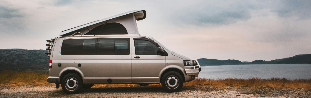 Silberner Campervan mit hochgeklapptem Dach steht an einem Seeufer mit bewlktem Himmel und Hgeln im Hintergrund.